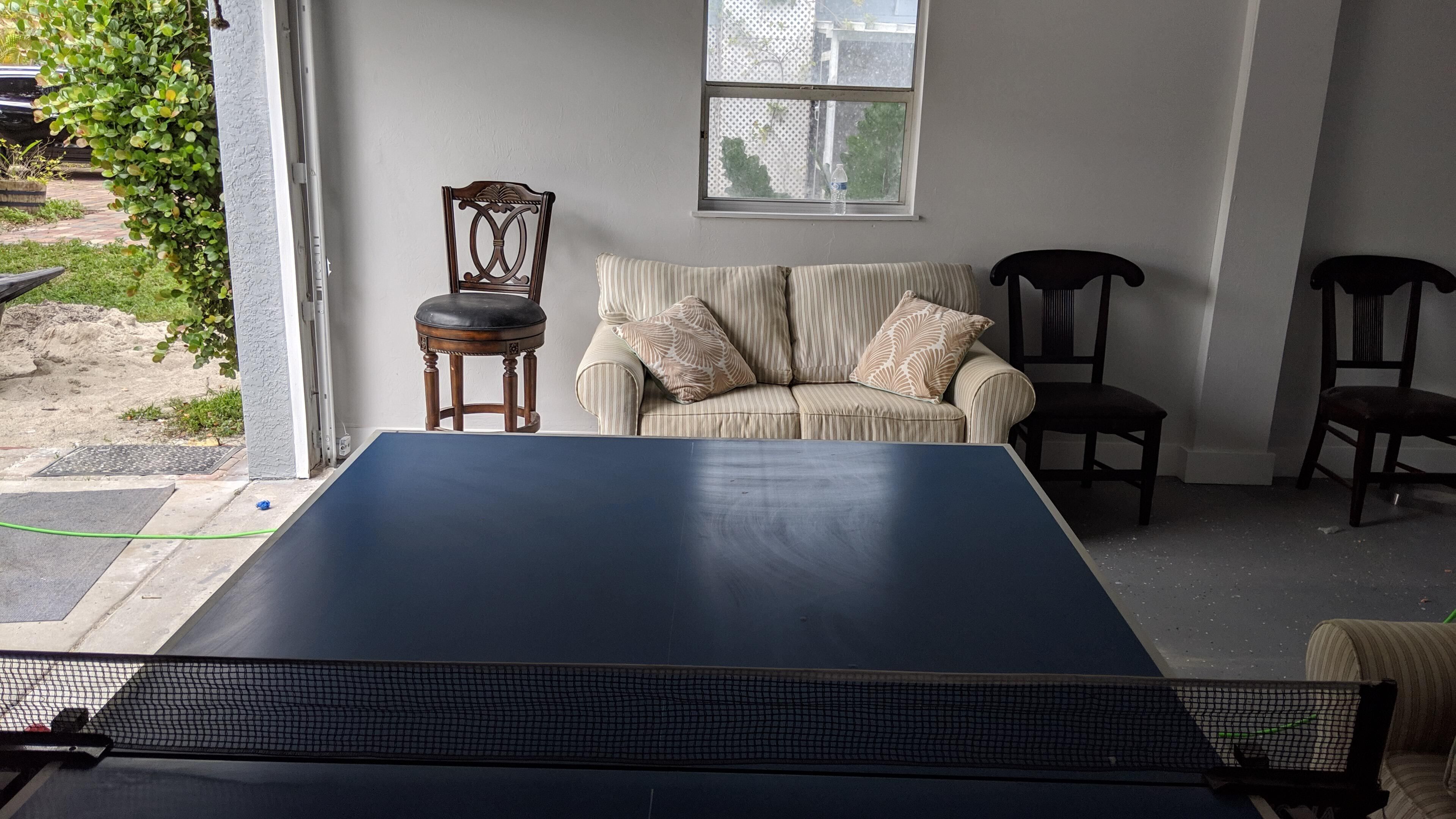 A ping pong table in a room with a sofa and two chairs, a window, and a view of greenery outside.
