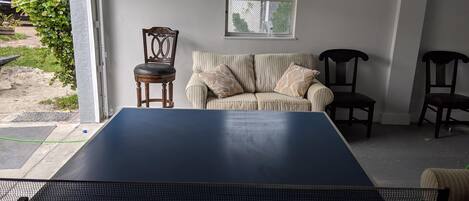 A ping pong table in a room with a sofa and two chairs, a window, and a view of greenery outside.