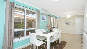 A dining area with a white table and chairs, large windows with curtains, and a view of the outside.