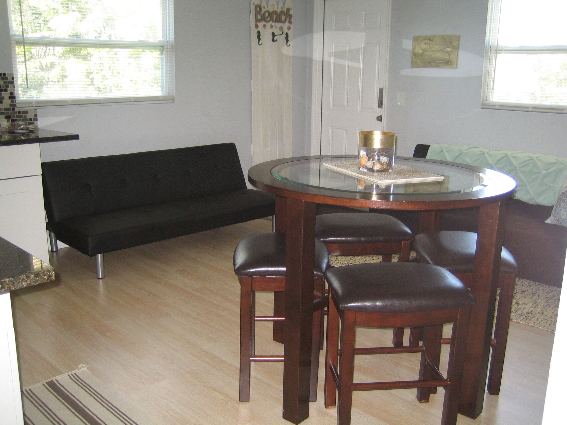 A compact living space with a round glass table surrounded by four chairs, a black sofa, and a door with a nameplate.