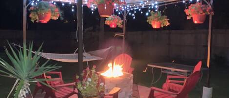 A covered outdoor seating area with a fire pit, red chairs, hanging flower pots, and string lights.