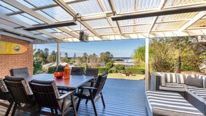 A covered outdoor dining area with a table and chairs, a cushioned seating area, and a view of the ocean and surrounding landscape. - Casa Moana - views of the beach (Gerringong)