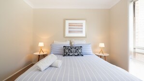 A neatly made bed with striped bedding, two bedside lamps, a framed picture above the bed, and a window with blinds on the right. - Casa Moana - views of the beach (Gerringong)
