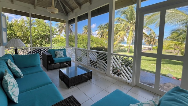 A covered outdoor seating area with a sofa and coffee table, large windows with a view of palm trees and greenery.