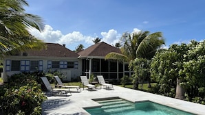 A poolside area with lounge chairs and a swimming pool, surrounded by lush greenery and a house with a tiled roof.