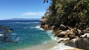 Una playa con agua azul cristalina, orilla arenosa y afloramientos rocosos.