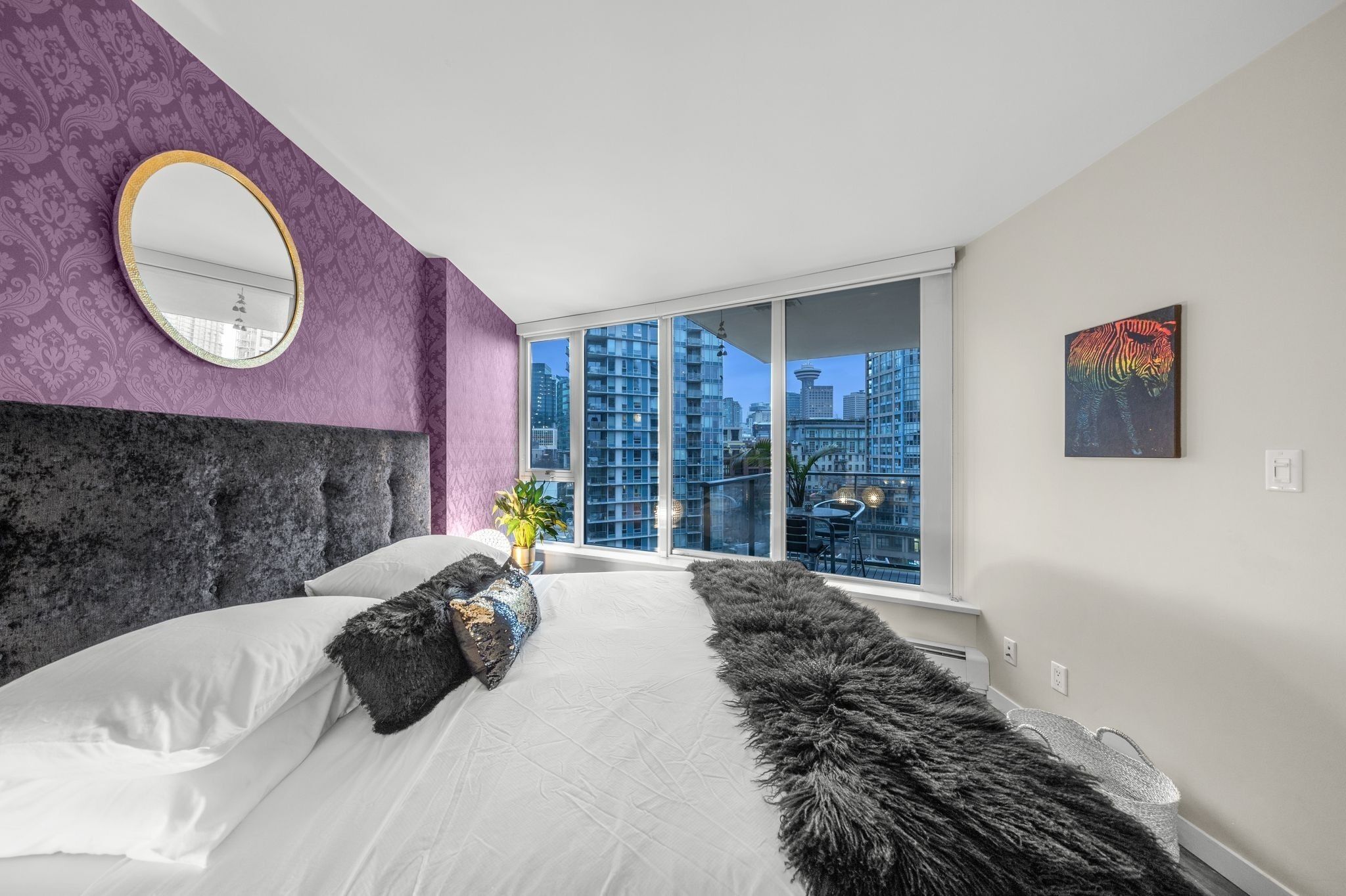 Une chambre moderne avec un grand lit, un miroir rond et une vue sur la ville grâce à de grandes fenêtres.