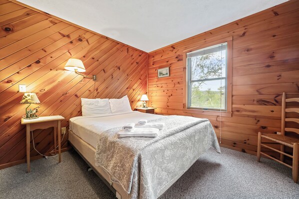 Ett sovrum med träpanel på väggarna, en säng, två sänglampor, ett fönster och ett litet bord med en lampa.