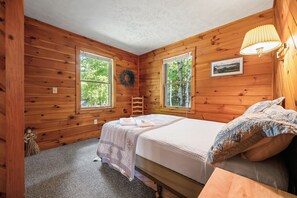 Een knus kamer in een blokhut met twee bedden, een houten wand, een raam met uitzicht op bomen, en een foto aan de muur.