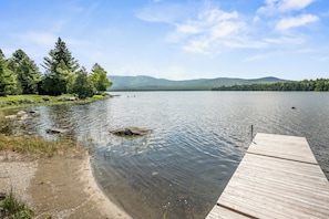 A calm lake with a wooden dock extending into the water, surrounded by dense forest and distant mountains.
