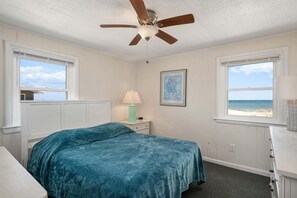A bedroom with a bed, a ceiling fan, a nightstand with a lamp, a framed picture, and a window with a beach view. - 405 Ocean-Front with Panoramic Ocean Views (Nags Head)