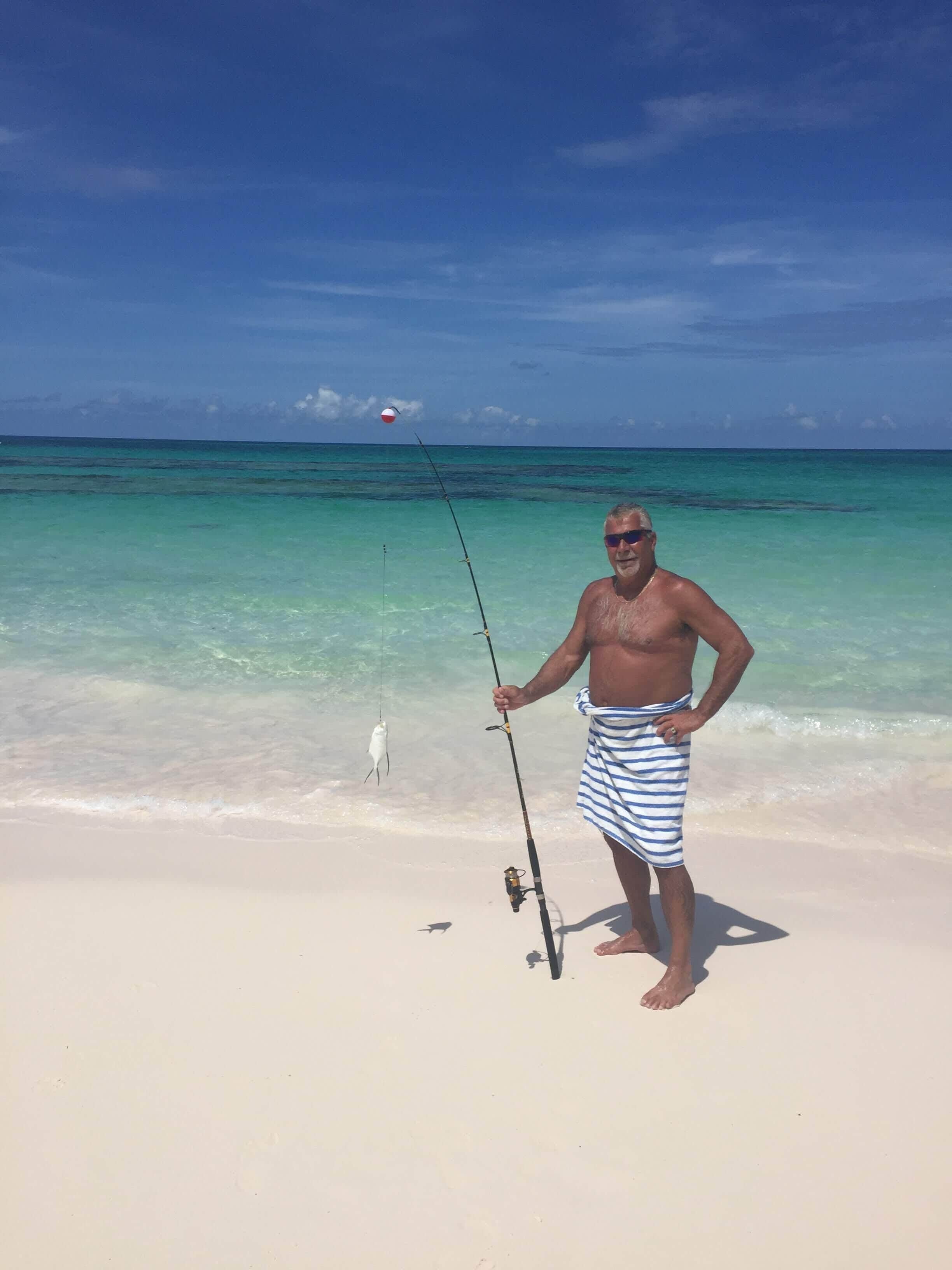 Un hombre de pie en la playa sosteniendo una caña de pescar con un pez.