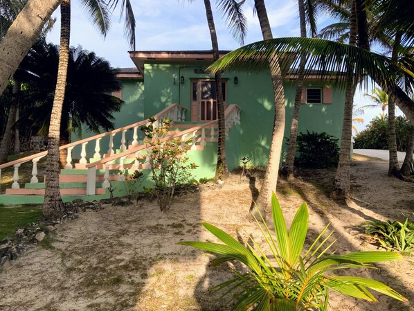 Un bâtiment vert avec un escalier menant à une porte, entouré de palmiers et bordé d’un sol sablonneux.