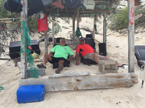 A beach scene with a wooden structure, a man in a green shirt, and another in a red shirt sitting under a shaded area.