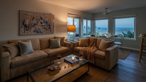 Sala de estar com um sofá bege, uma mesa de centro de vidro e um quadro na parede. Há vista para o oceano através de janelas amplas.