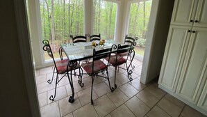 A dining area with a glass table and chairs, large windows with a forest view, and a ceiling light fixture.