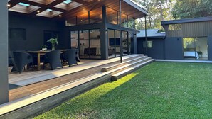 A modern house with a covered patio, a dining area, and a kitchen visible through glass doors. - Private Architectual Beach Retreat in Pearl Beach (pearl beach)