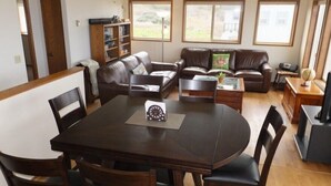 A living room with a dark wooden dining table, leather sofas, and a bookshelf. - Dunes House at Bandon - 3 bedroom Oceanfront (Bandon)