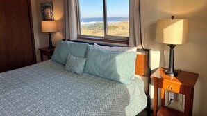 A bedroom with a bed, a window with a view of the beach, a lamp, and a painting on the wall. - Dunes House at Bandon - 3 bedroom Oceanfront (Bandon)