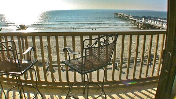 Un balcon avec des chaises et une table en métal, donnant sur une plage et une jetée.