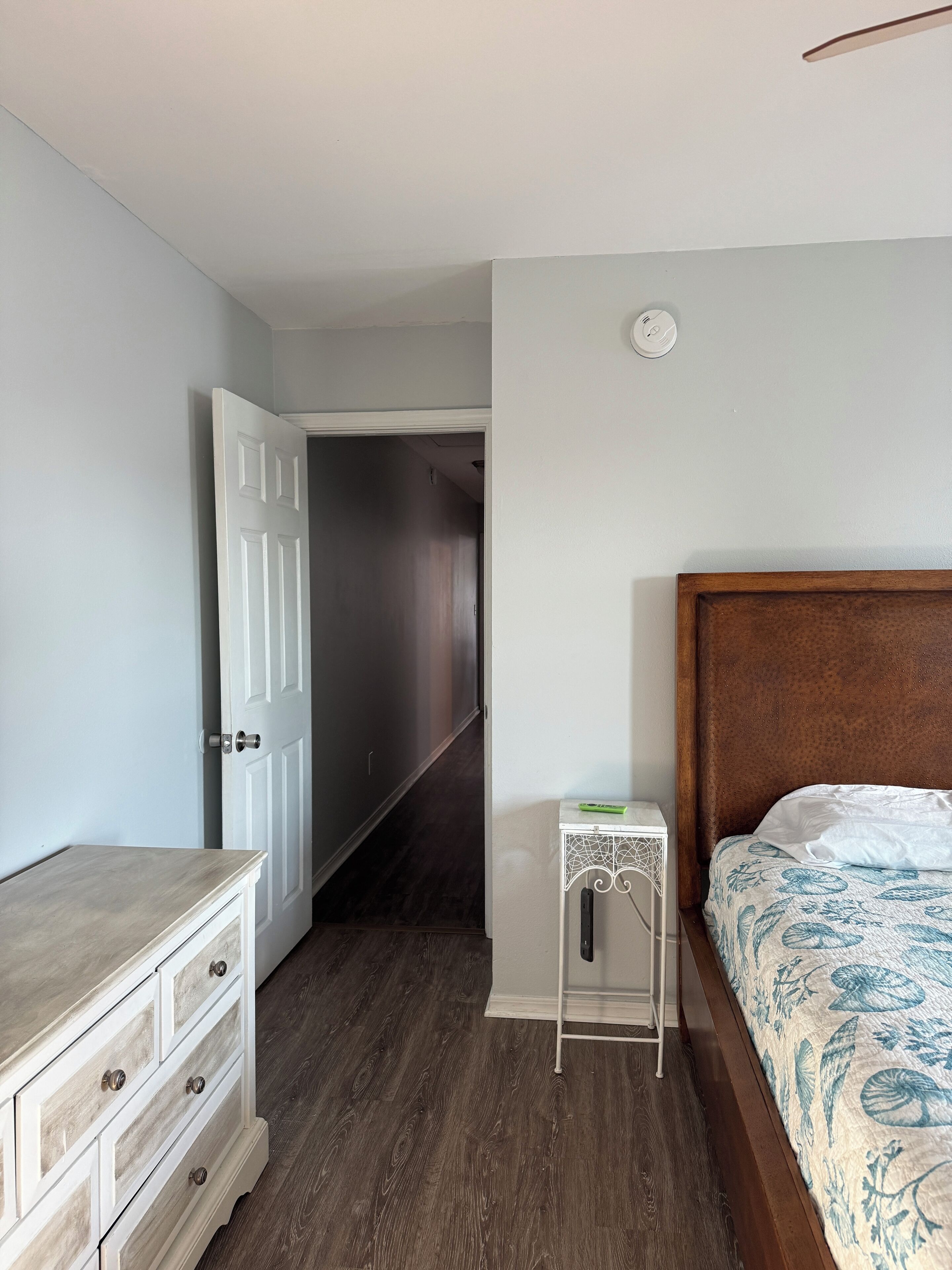 A bedroom with a wooden dresser, a bedside table, a bed with a patterned bedspread, and a door leading to another room.