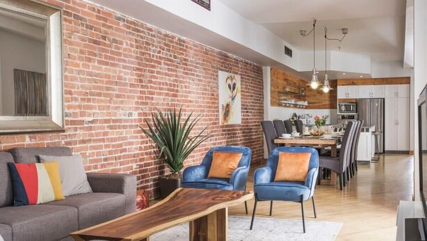 Ein modernes Wohnzimmer mit Backsteinwand, einem Holztisch, grauen Sofas, blauen Sesseln und einem Essbereich mit Küche im Hintergrund.