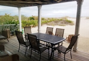 Ein überdachter Außenbereich mit Tisch und Stühlen mit Blick auf einen Sandstrand und Dünen.