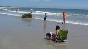 A beach with people walking and playing, a large rock in the water, and a green beach chair.