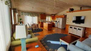A spacious living area with a wooden ceiling, a kitchen in the background, a sofa, a coffee table, and a television mounted on the wall.