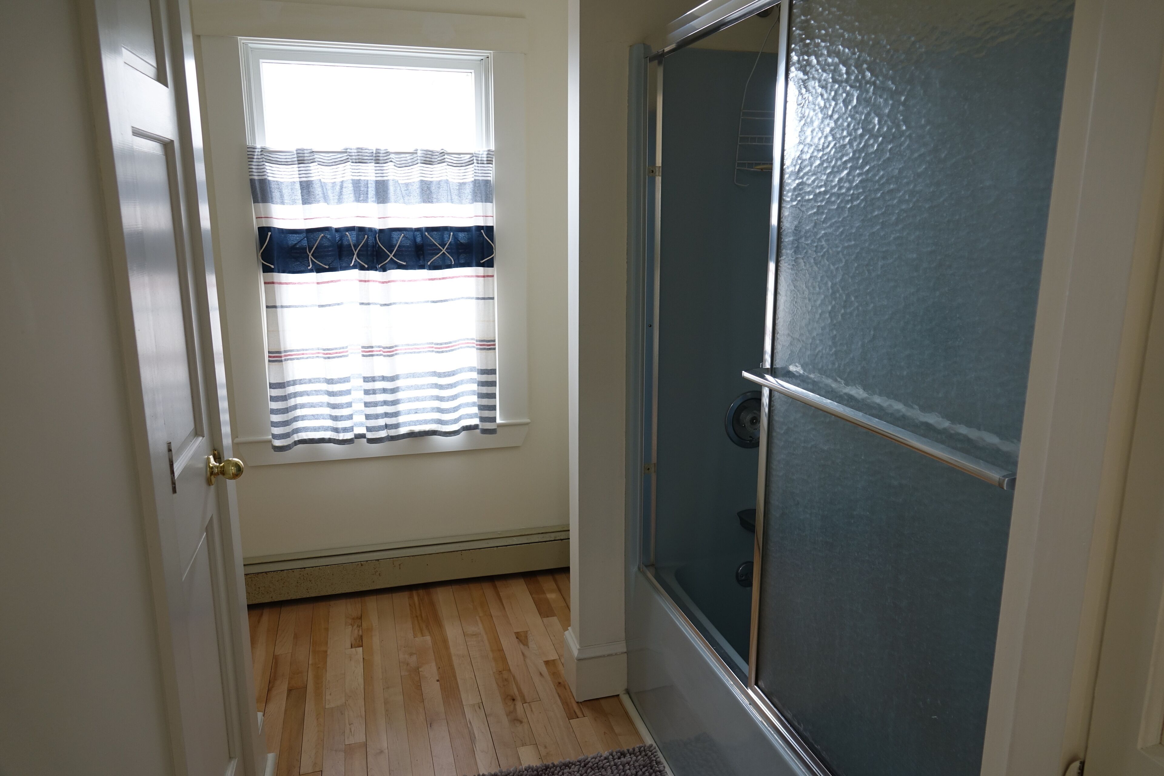 Een kamer met een houten vloer, een deur met een messing handgreep, een raam met gestreepte gordijnen en een douche met een glazen deur.