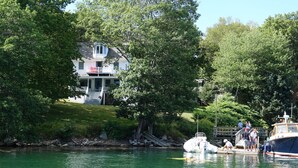 A house with a porch and steps leading to a deck, surrounded by trees and a body of water. - Waterfront Cottage in South Bristol w/ private dock and lobster boat views (South Bristol)