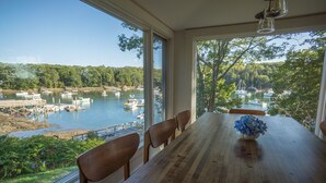 A dining area with a wooden table and chairs, overlooking a lake with boats and docks. - Waterfront Cottage in South Bristol w/ private dock and lobster boat views (South Bristol)