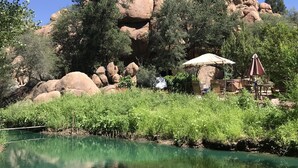 Et naturligt landskab med et klart vandareal, store klippeformationer og et siddeområde med en parasol.