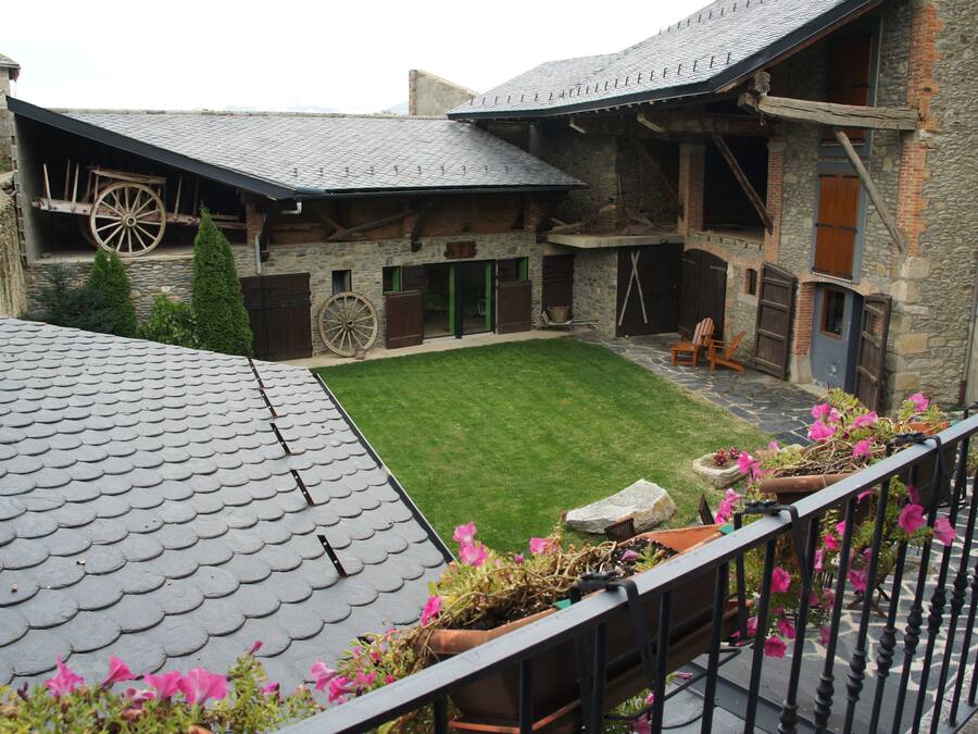 A stone building with a sloped roof, a wooden wheel, and a fenced courtyard with potted plants.