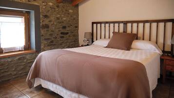 Une chambre avec un mur en pierre, un plafond en bois, un lit recouvert d’une couverture marron, une table de chevet avec une lampe et une fenêtre avec un rideau.