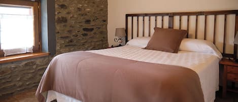 Un dormitorio con pared de piedra, techo de madera, cama con colcha marrón, mesita de noche con lámpara y ventana con cortina.