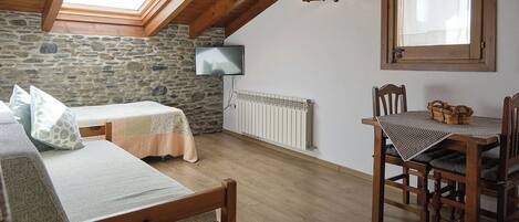 Ein gemütliches Zimmer mit Steinwand, Holzbalkendecke, einem Bett, einem Tisch mit Stühlen und einem an der Wand montierten Fernseher.