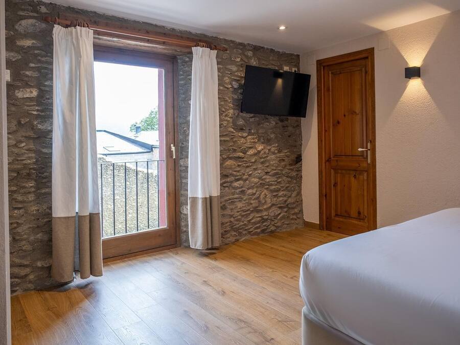 Un dormitorio con pared de piedra, puerta de madera, televisor montado en la pared y balcón con cortinas.
