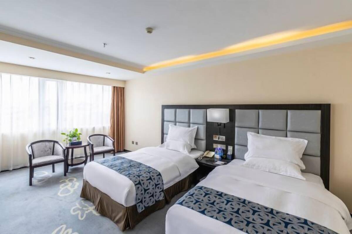 A hotel room with two beds, a nightstand, a lamp, and a window with curtains.