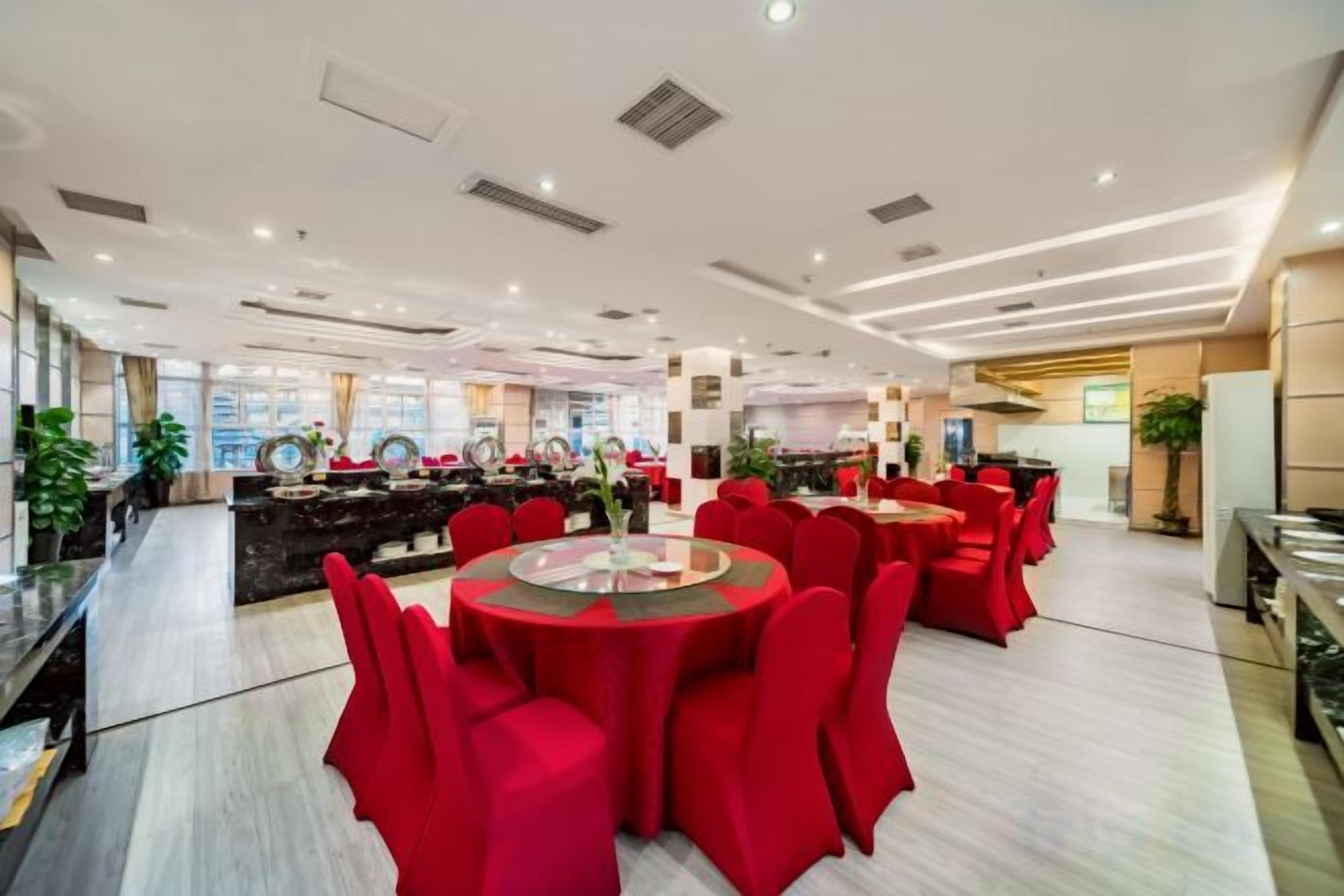 A spacious hotel dining area with round tables and red chairs, a reception desk, and a view of the city.