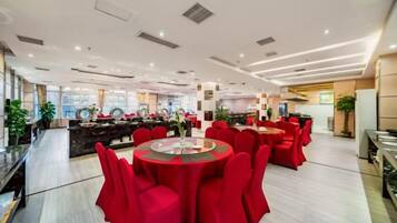 A spacious hotel dining area with round tables and red chairs, a reception desk, and a view of the city.