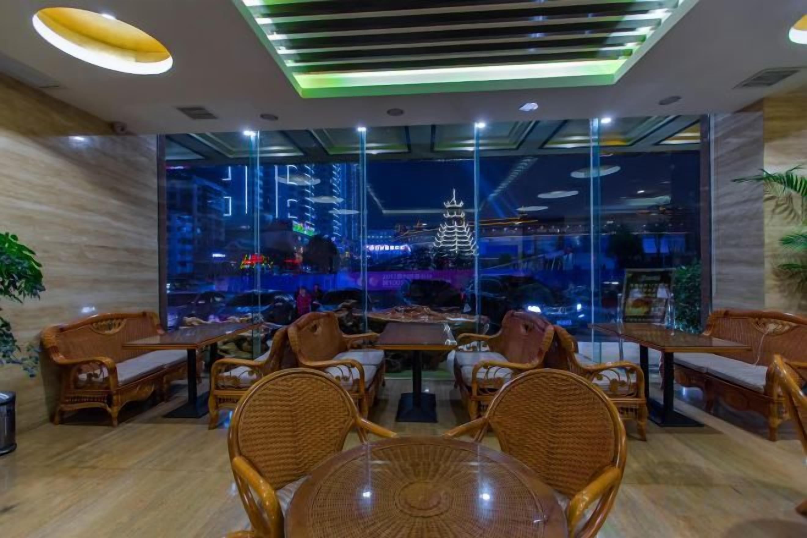 A modern hotel lobby with wicker furniture, a glass wall with a city view, and a ceiling with recessed lighting.