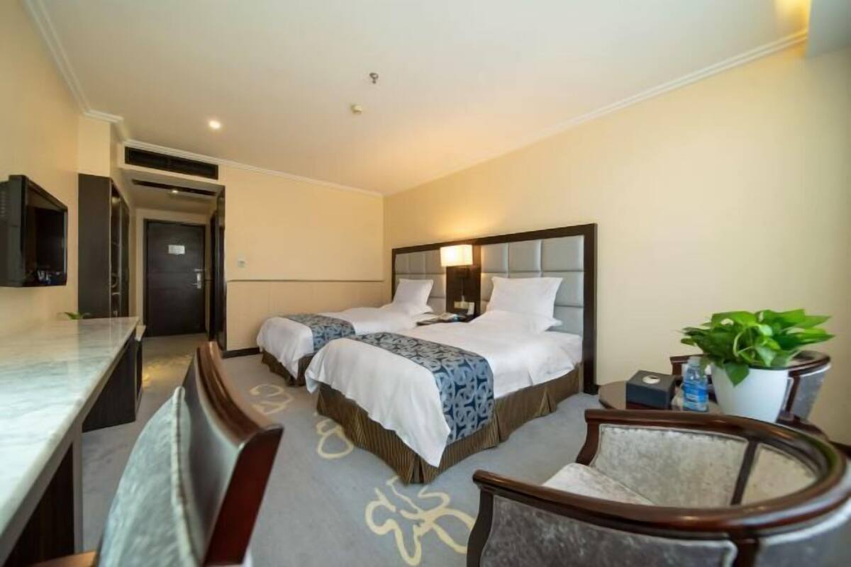 A hotel room with two beds, a desk, a chair, a potted plant, and a television.