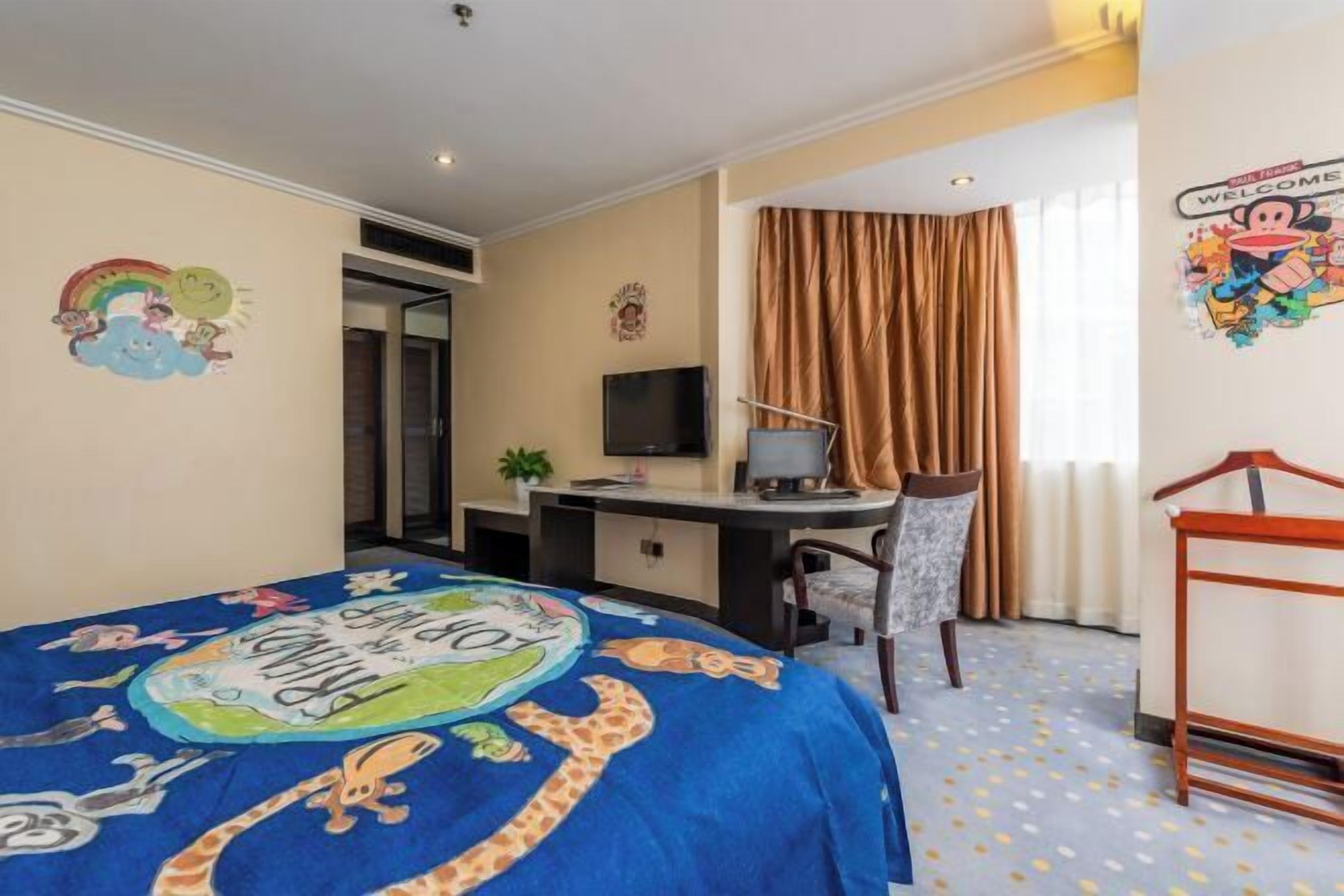Ein Hotelzimmer mit einem Bett, das mit einer farbenfrohen Decke bezogen ist, einem Schreibtisch mit einem Computer, einem Fernseher und einem Fenster mit Vorhängen.