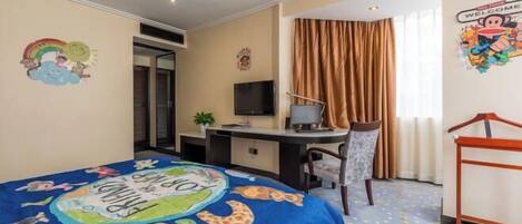 Ein Hotelzimmer mit einem Bett, das mit einer farbenfrohen Decke bezogen ist, einem Schreibtisch mit einem Computer, einem Fernseher und einem Fenster mit Vorhängen.