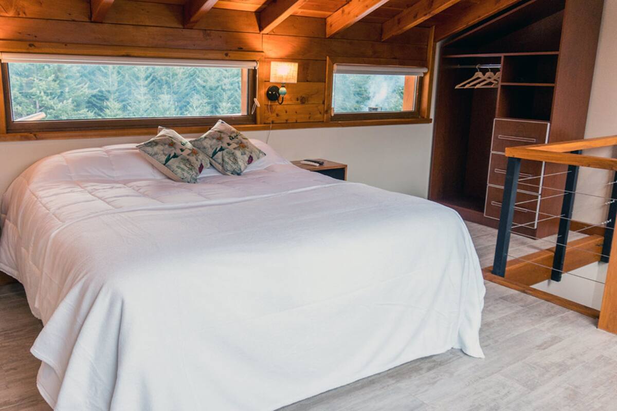 Ein Schlafzimmer mit einem großen Bett, zwei Nachttischen, einem Kleiderständer und einem Fenster mit Blick auf Bäume.