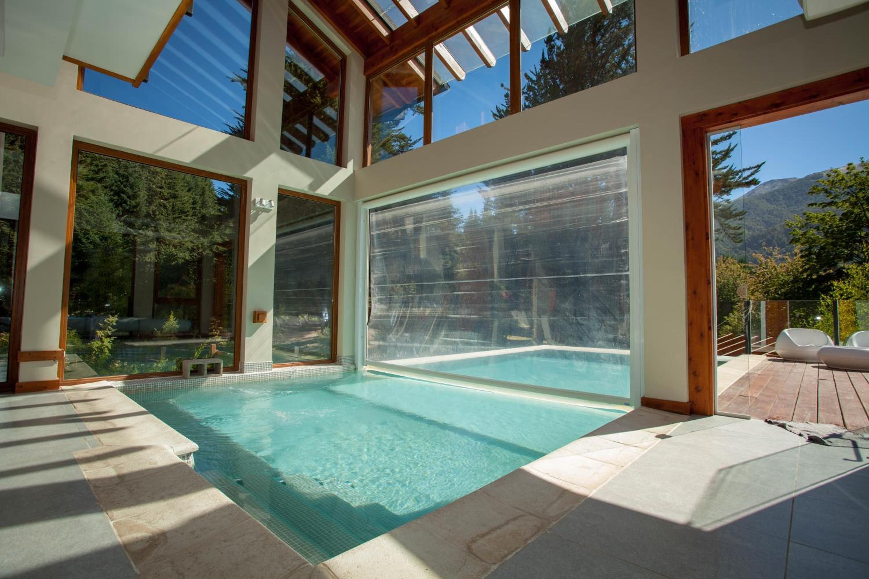 Ein moderner, überdachter Swimmingpool mit großen Glasüren, einem Holzboden und Blick auf Bäume und Berge.