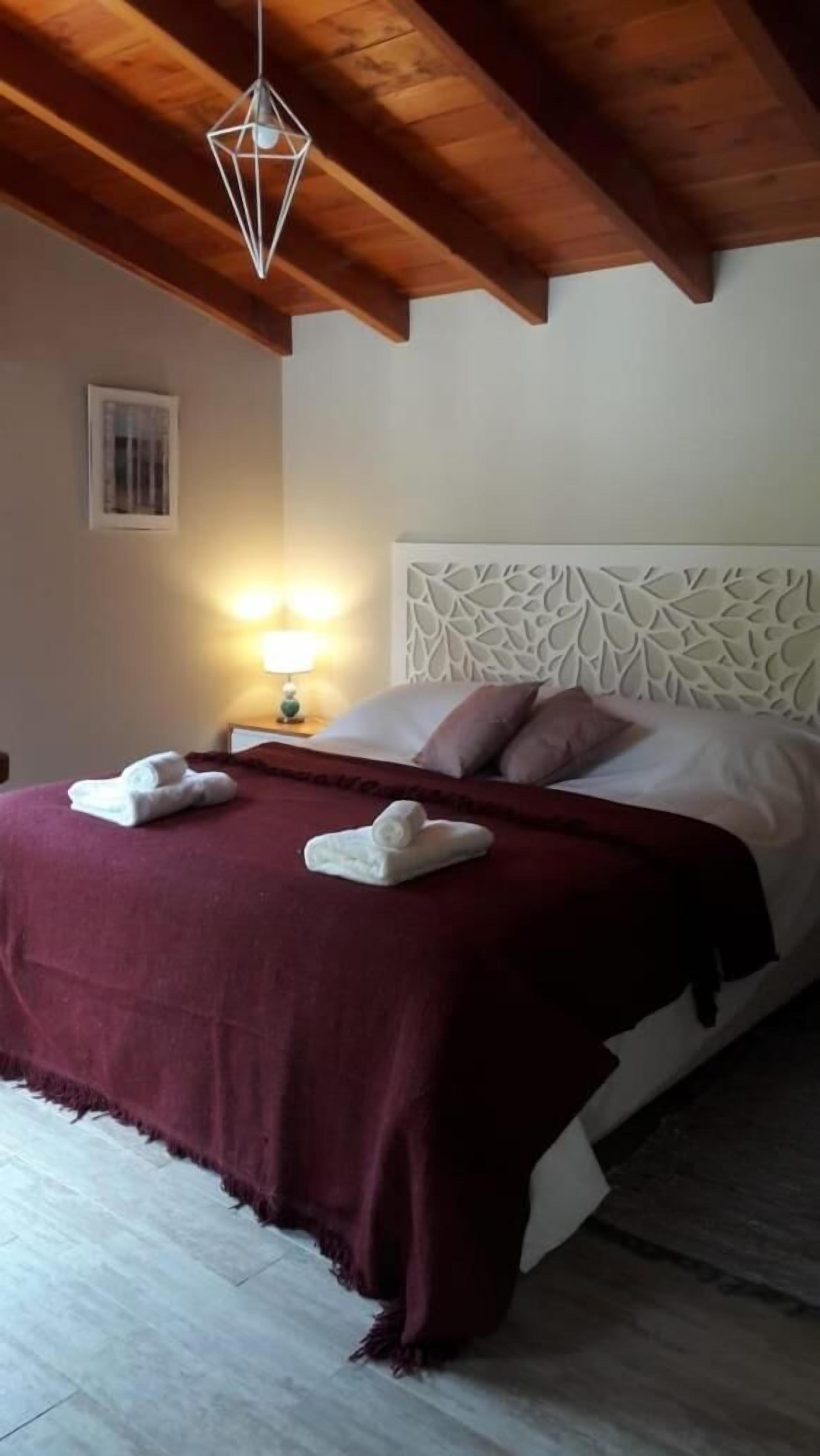 A hotel room with a wooden ceiling, a bed with a white headboard, a red blanket, and two towels on the bed.