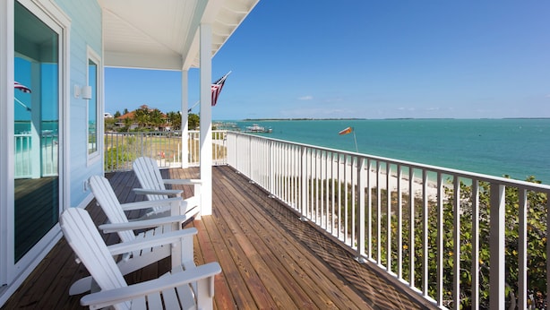 A wooden deck with white chairs, a view of the ocean, and a flag. - The Blue Marlin - Beachfront Oasis with Private Beachside Pool & Boat Dock (Captiva)