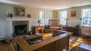 A traditional living room with a fireplace, wooden furniture, and a view of the outdoors through large windows. - Large, Charming Cape Cod Home Close To Orleans And Chatham Beaches (Orleans)